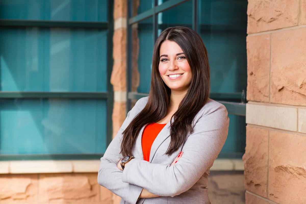 A confident woman with long brown hair, wearing a gray blazer and red top, standing with arms crossed outdoors near a brick wall and windows.