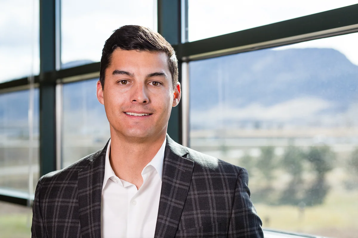 Portrait of a young man with dark hair in a checkered suit and white shirt, standing indoors near large windows with mountain scenery outside.