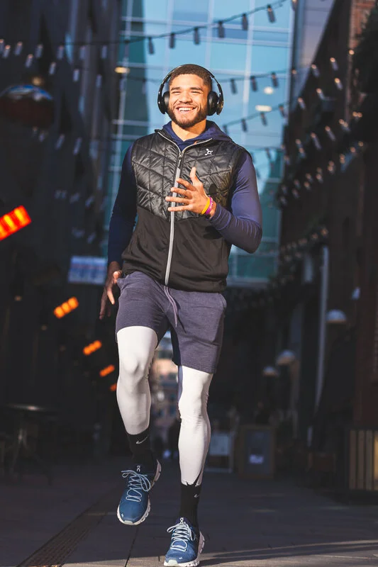 A man jogging outdoors wearing headphones, a black vest, long sleeve shirt, gray shorts, and wearing compression leggings and athletic shoes, smiling and enjoying his run.