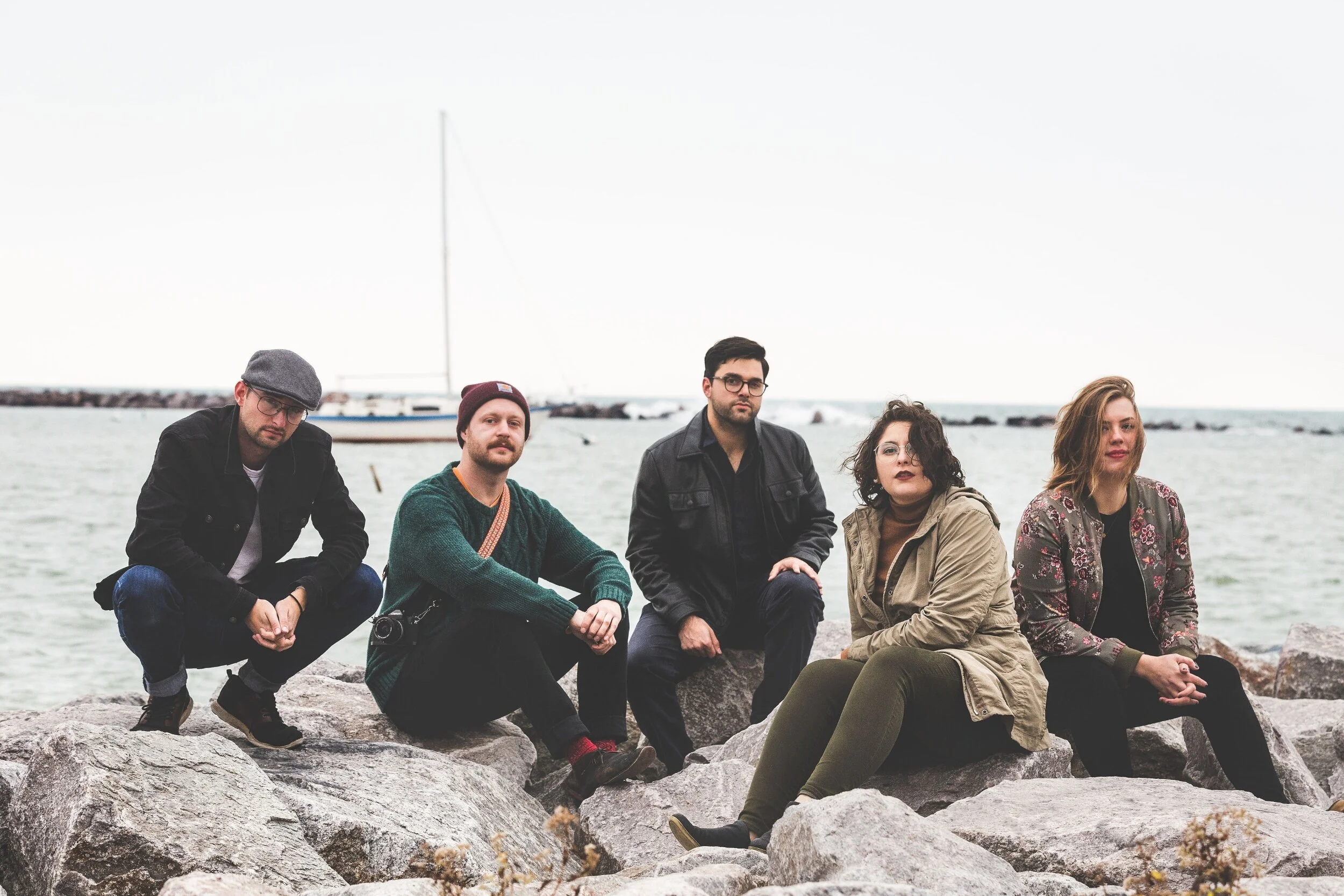Five people sitting on rocks near the water, with a boat and sailboat in the background on an overcast day.