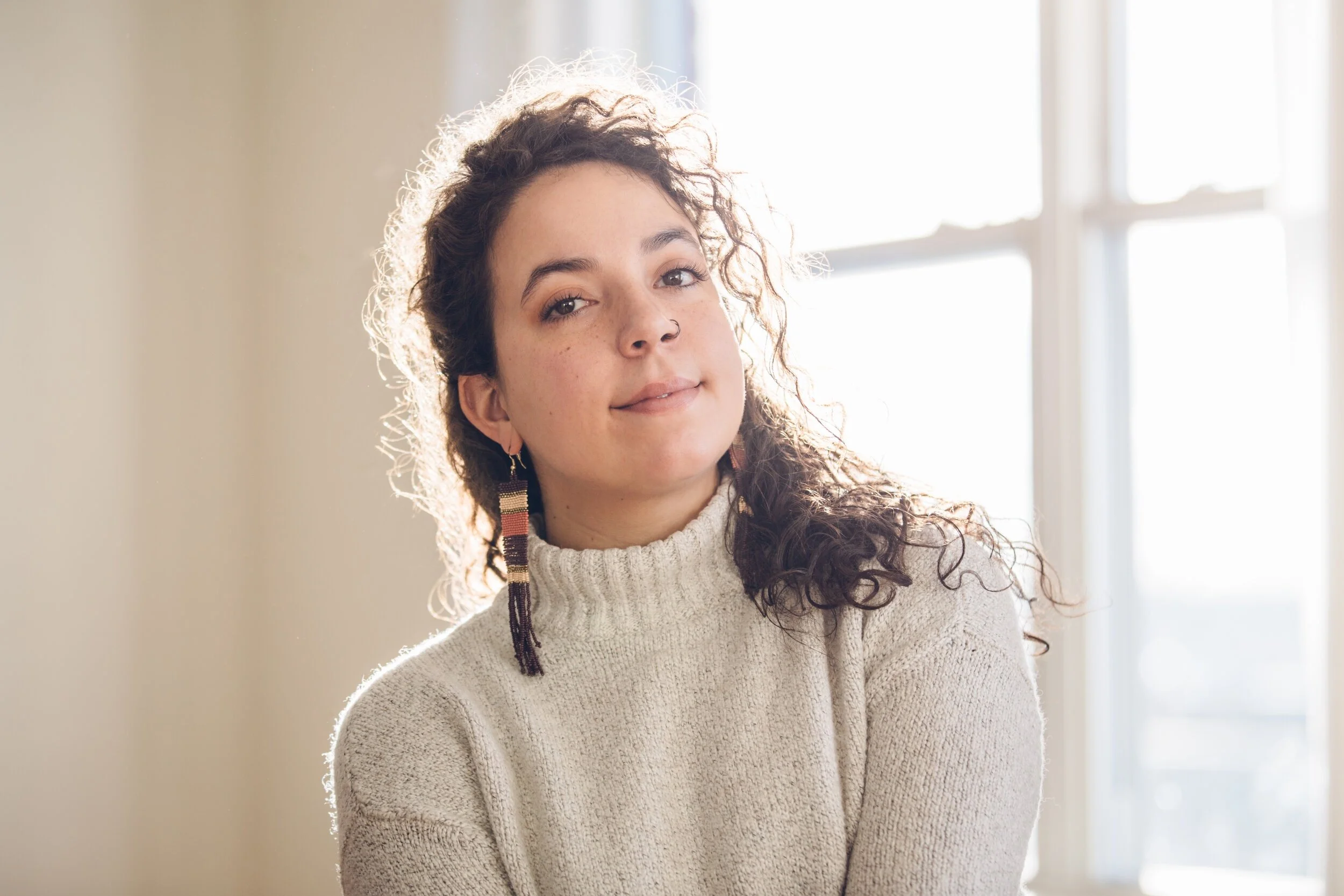 A woman with curly dark hair and light skin, wearing a beige sweater and colorful earrings, sitting near a window with sunlight behind her.