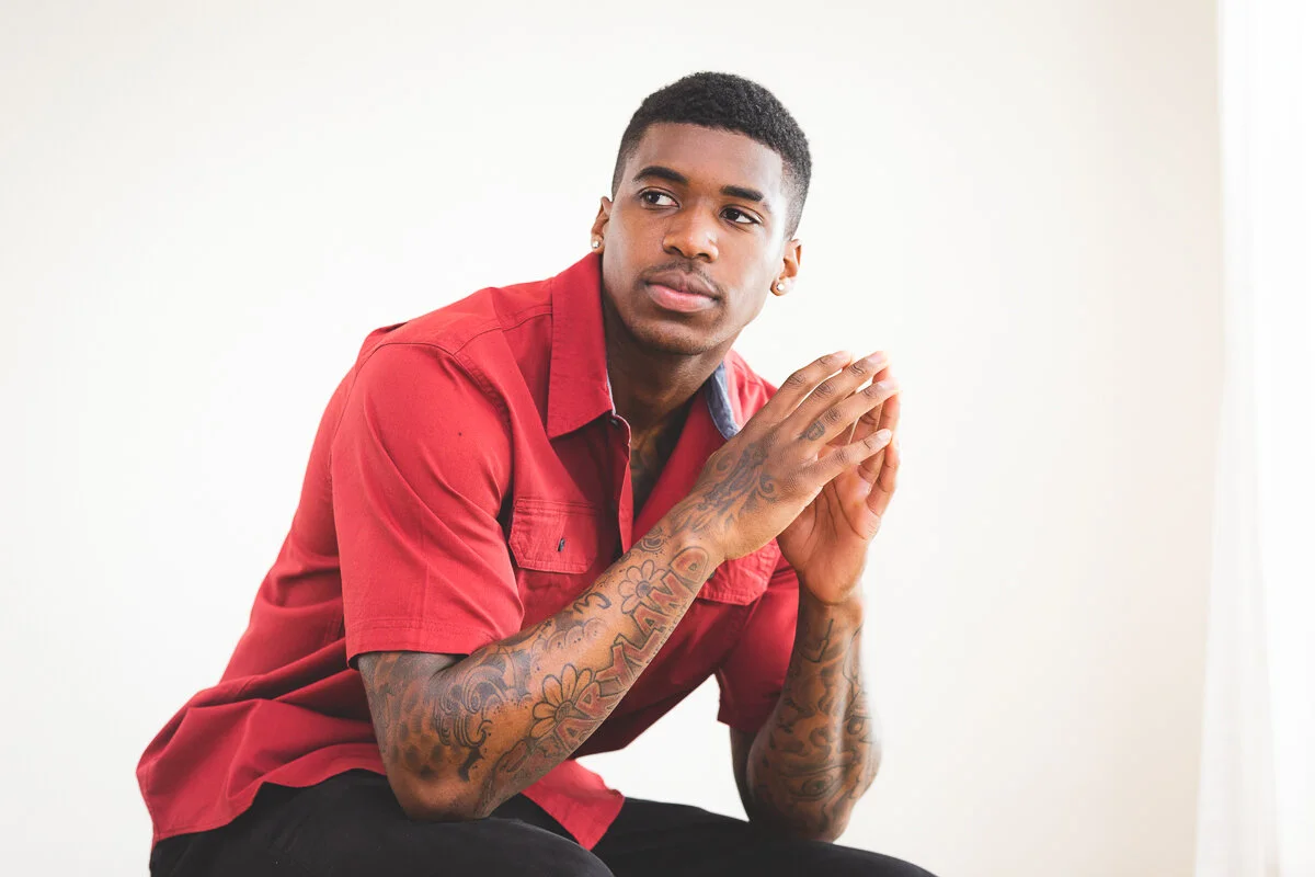 A young man with short black hair, wearing a red shirt and black pants, is sitting with his hands clasped together in front of his face, looking contemplative against a plain white background.