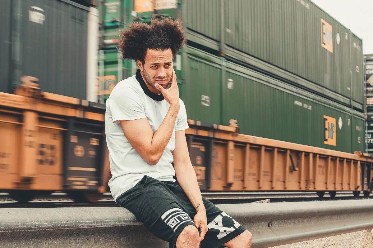 A man with curly hair sitting on a guardrail near train tracks, looking worried or confused. There are stacked green and orange freight containers moving in the background.