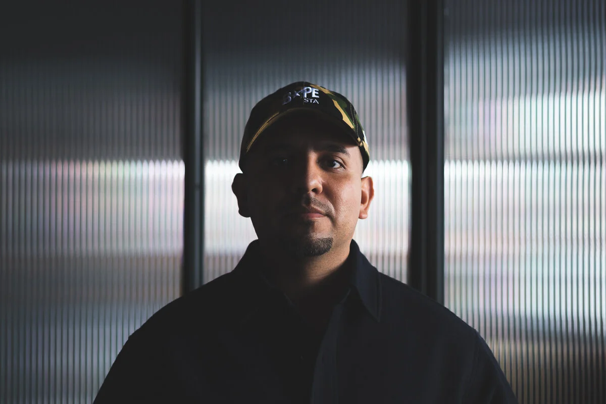 A man wearing a camouflage cap and black jacket standing in front of a metallic, ribbed background.