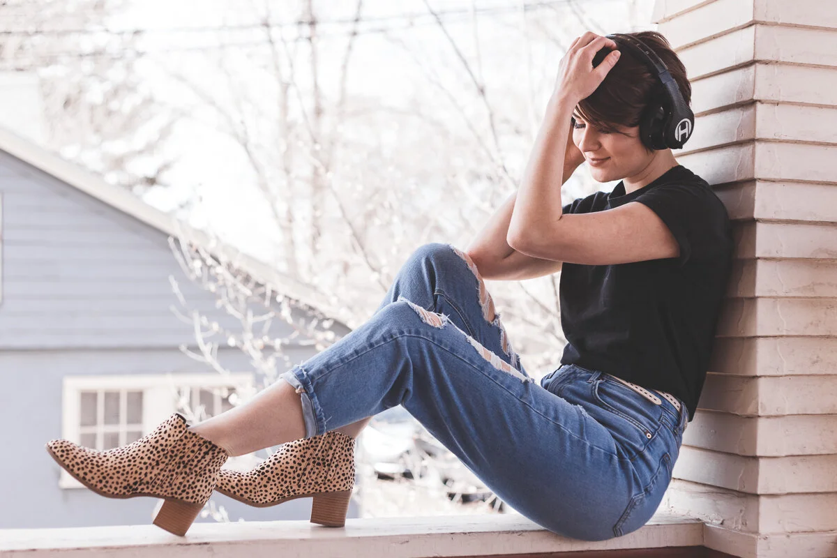 A woman sitting on a porch, wearing ripped jeans, animal print ankle boots, a black t-shirt, and headphones, leaning against a brick wall with trees and a house in the background.