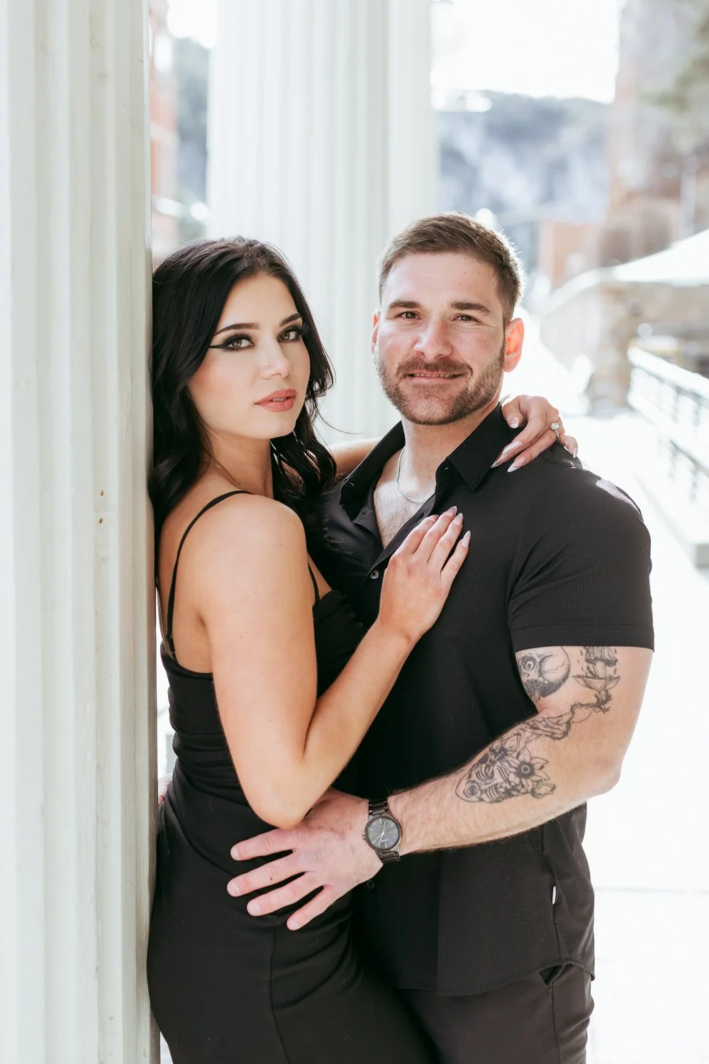 A woman with dark hair and makeup and a man with a beard and tattoos on his arm posing outdoors, both wearing black clothing, standing close to each other with the woman leaning against a wall.
