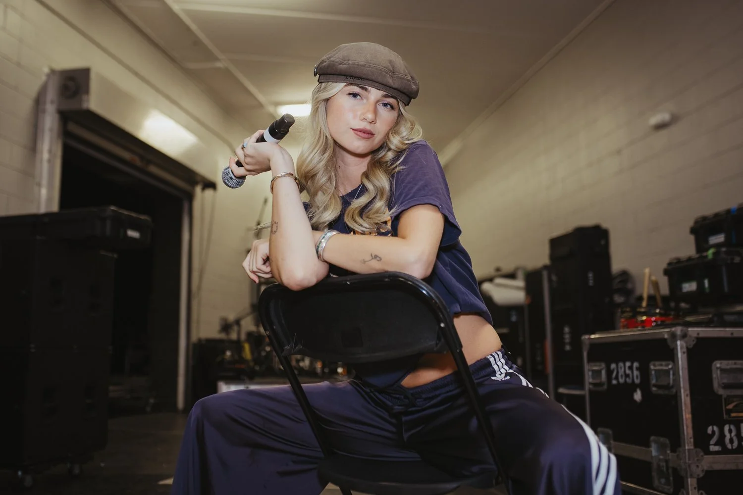 A young woman with blonde hair and tattoos, wearing a brown hat and a loose purple shirt, sitting on a black chair in a music rehearsal space, holding a microphone.
