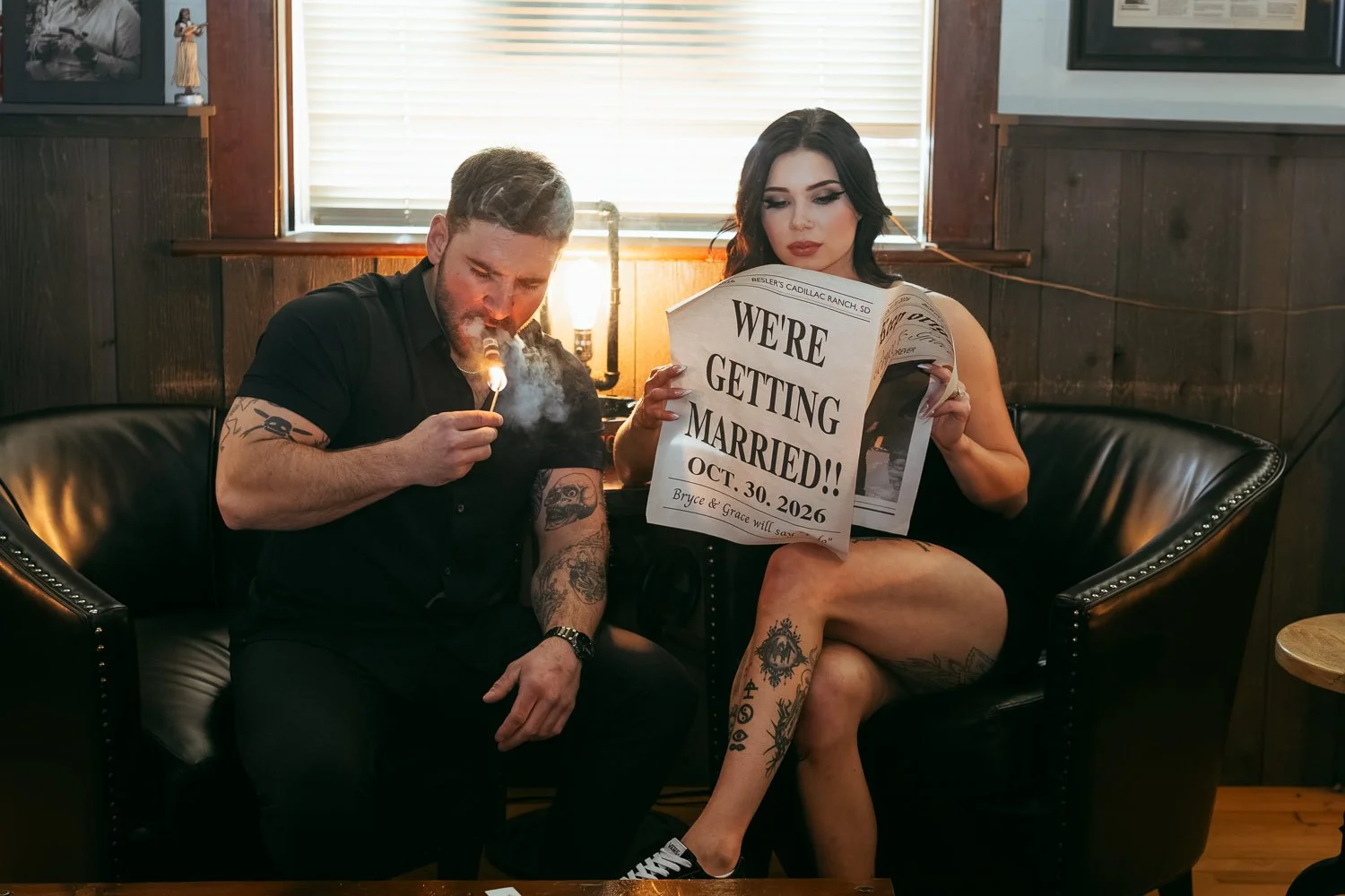 A man and woman sitting on a black leather couch, the woman holding a newspaper with 'We're getting married!!' written on it, and the man lighting a cigarette with a match.