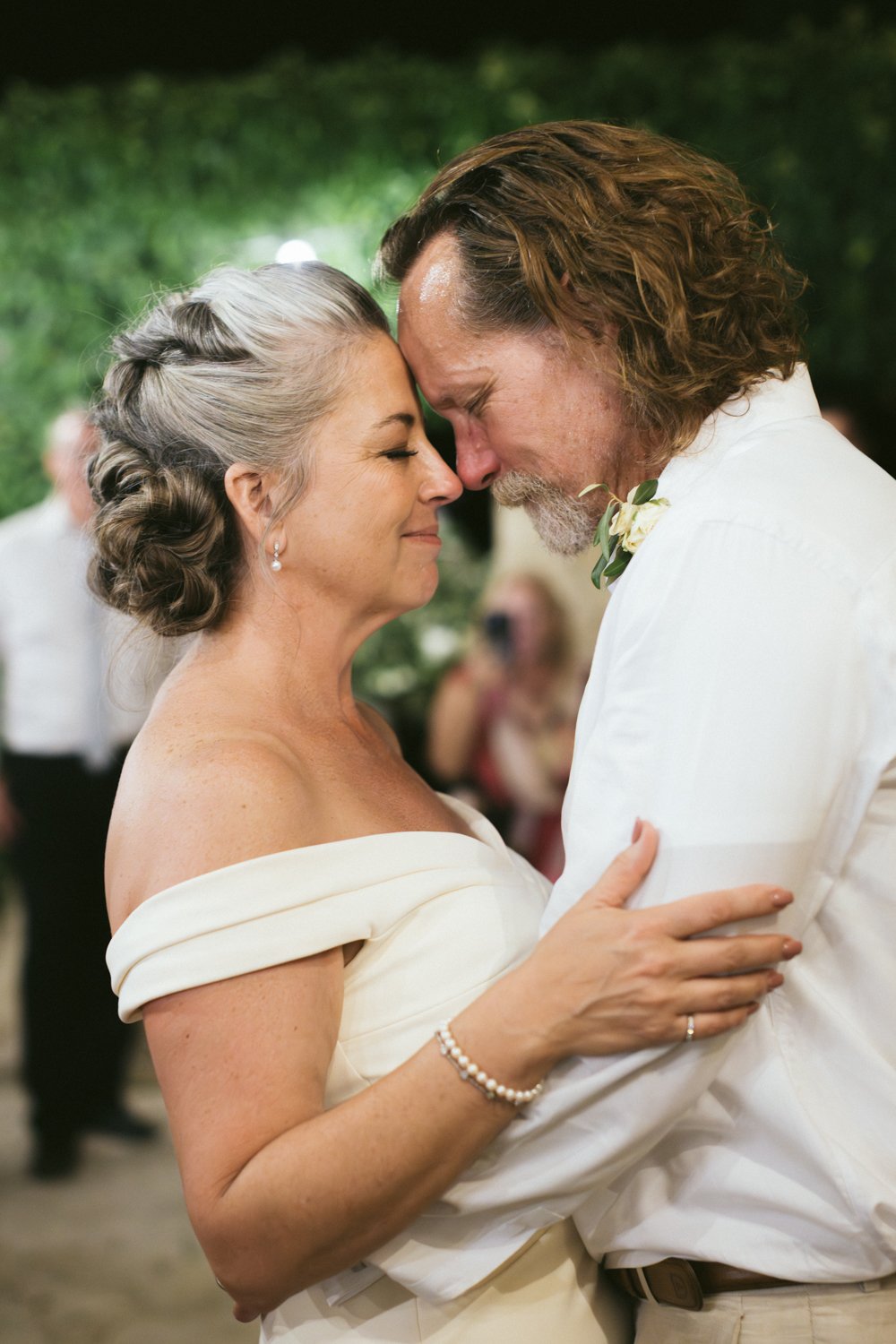 A middle-aged woman and an older man are dancing closely, touching foreheads and smiling with eyes closed at a wedding reception. The woman has silver hair styled in an elegant updo, and the man has long, curly brown hair and a beard. They are both d