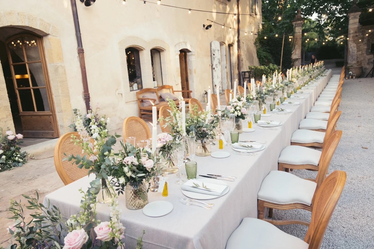 Long outdoor banquet table decorated with floral arrangements, candles, and tableware, set up for an event in a rustic courtyard with string lights.