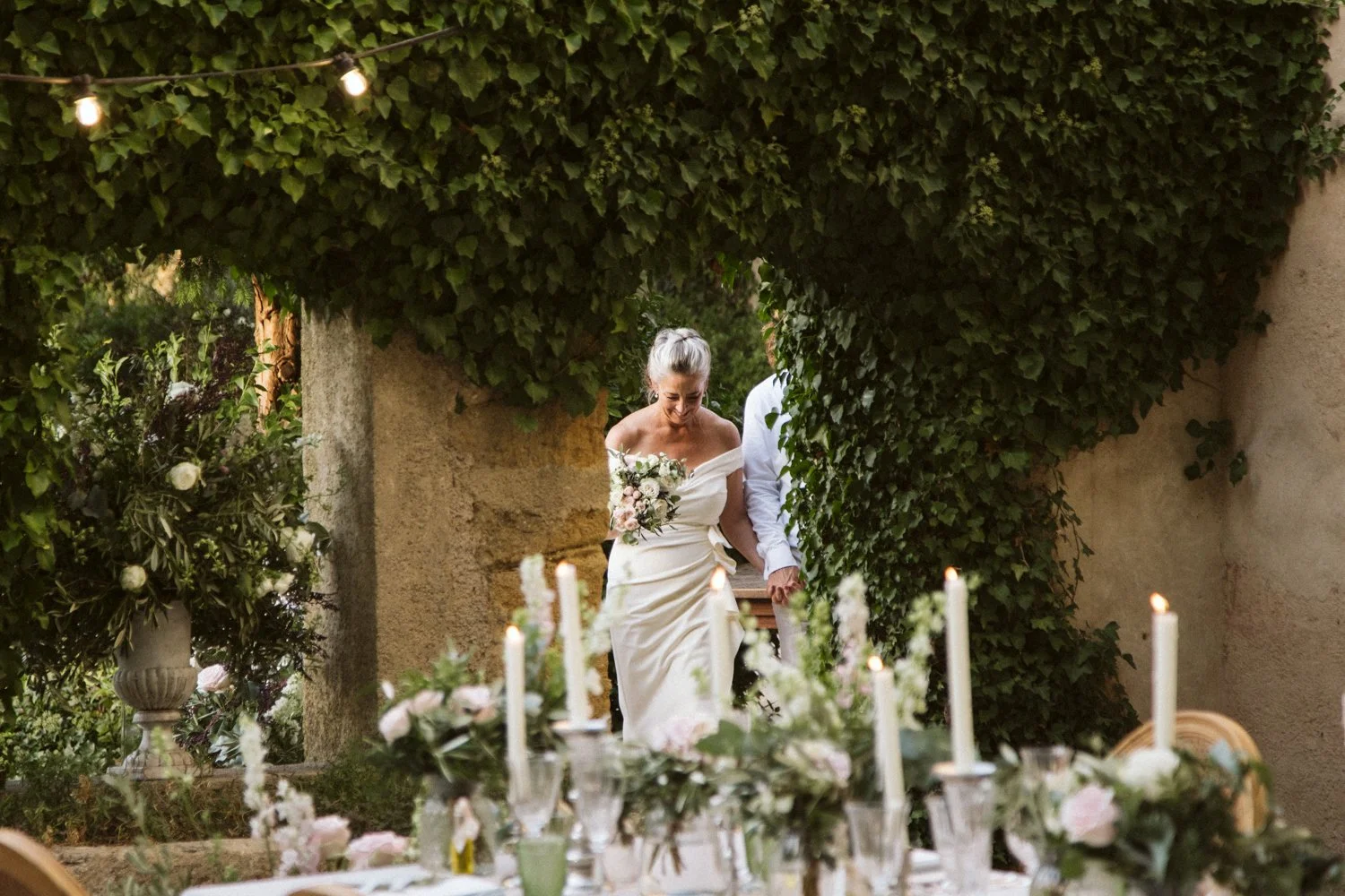 A bride and groom walking together in an outdoor setting decorated with greenery and candles, with a lush green ivy-covered wall and a table with floral arrangements and lit candles in the foreground.