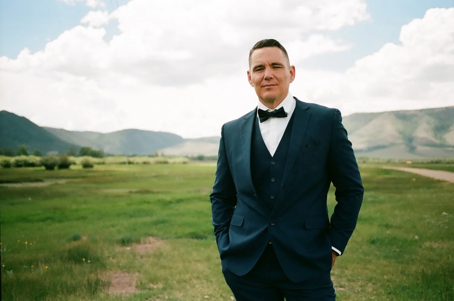 A man in a navy blue suit with a bow tie stands outdoors in a green field with mountains and a cloudy sky in the background.