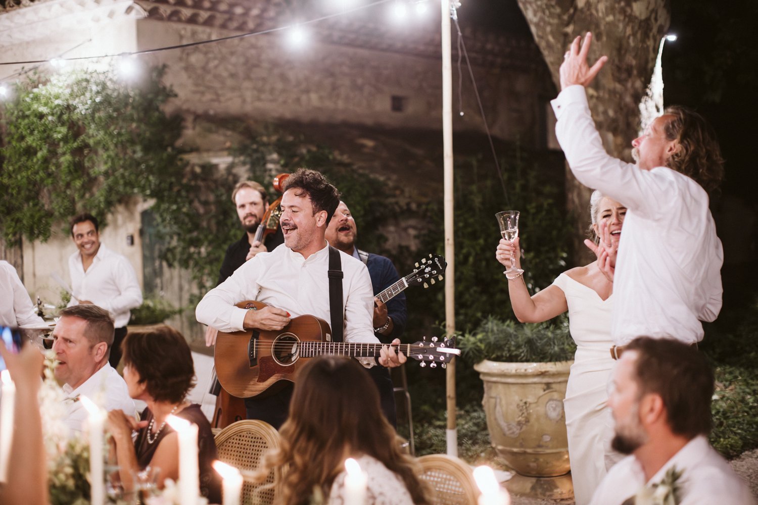 A lively outdoor celebration at night with musicians playing guitars and a woman holding a glass of wine, while guests enjoy and dance.