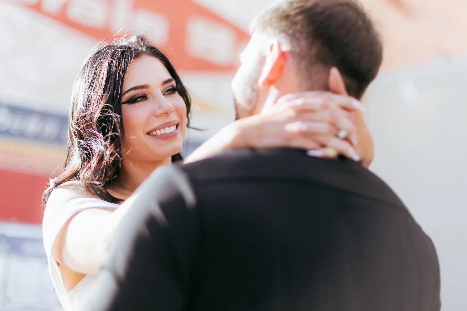 A woman with dark hair and makeup smiling, holding the face of a man with light brown hair, who is facing away from the camera.