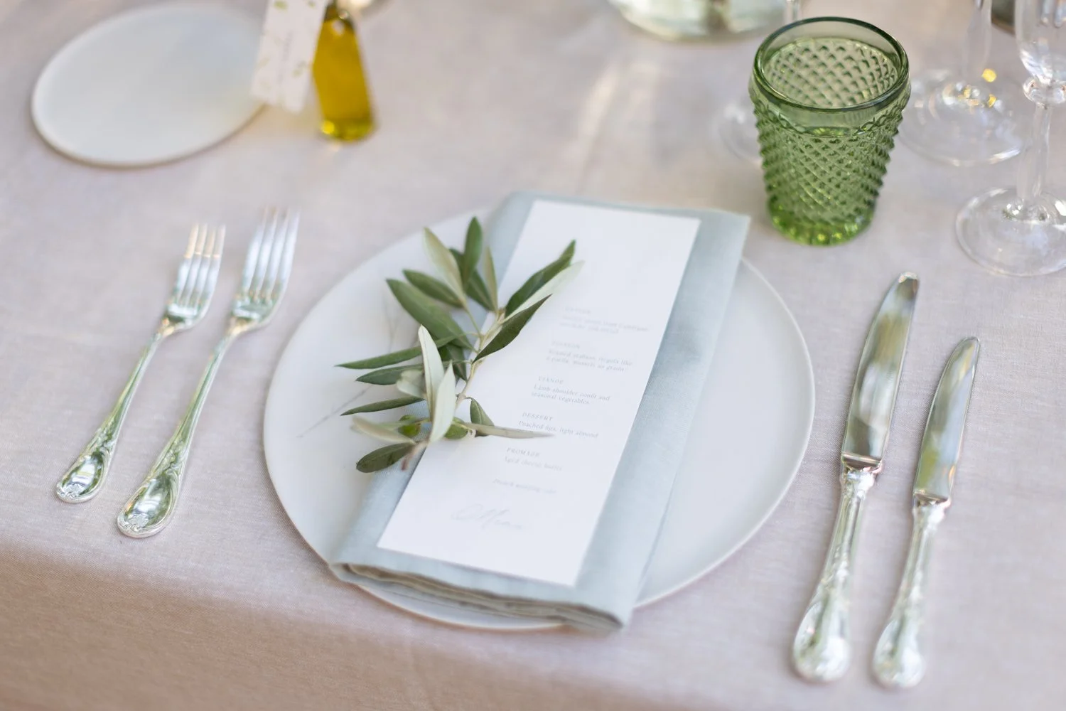 Elegant table setting with a white plate, silverware, a green glass, a menu with a sprig of greenery, and a light-colored tablecloth.