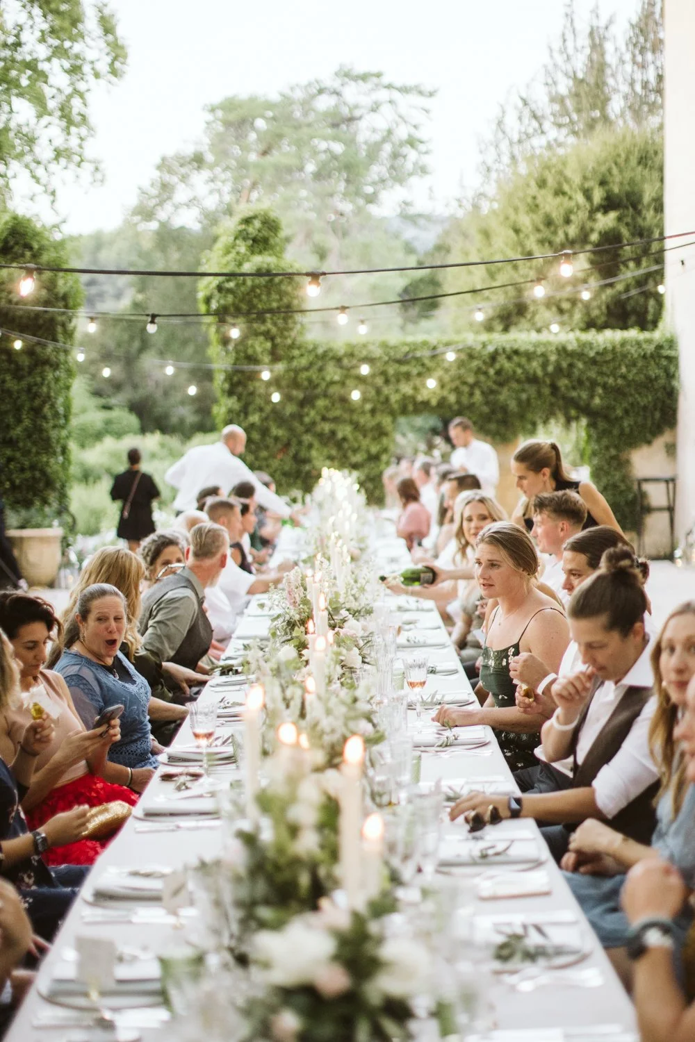 A long outdoor dining table with guests, floral centerpieces, and candles under hanging string lights in a garden setting.