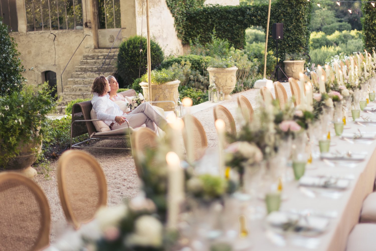 A wedding reception outdoors with a long decorated table and a bride and groom sitting on a bench in a garden setting.