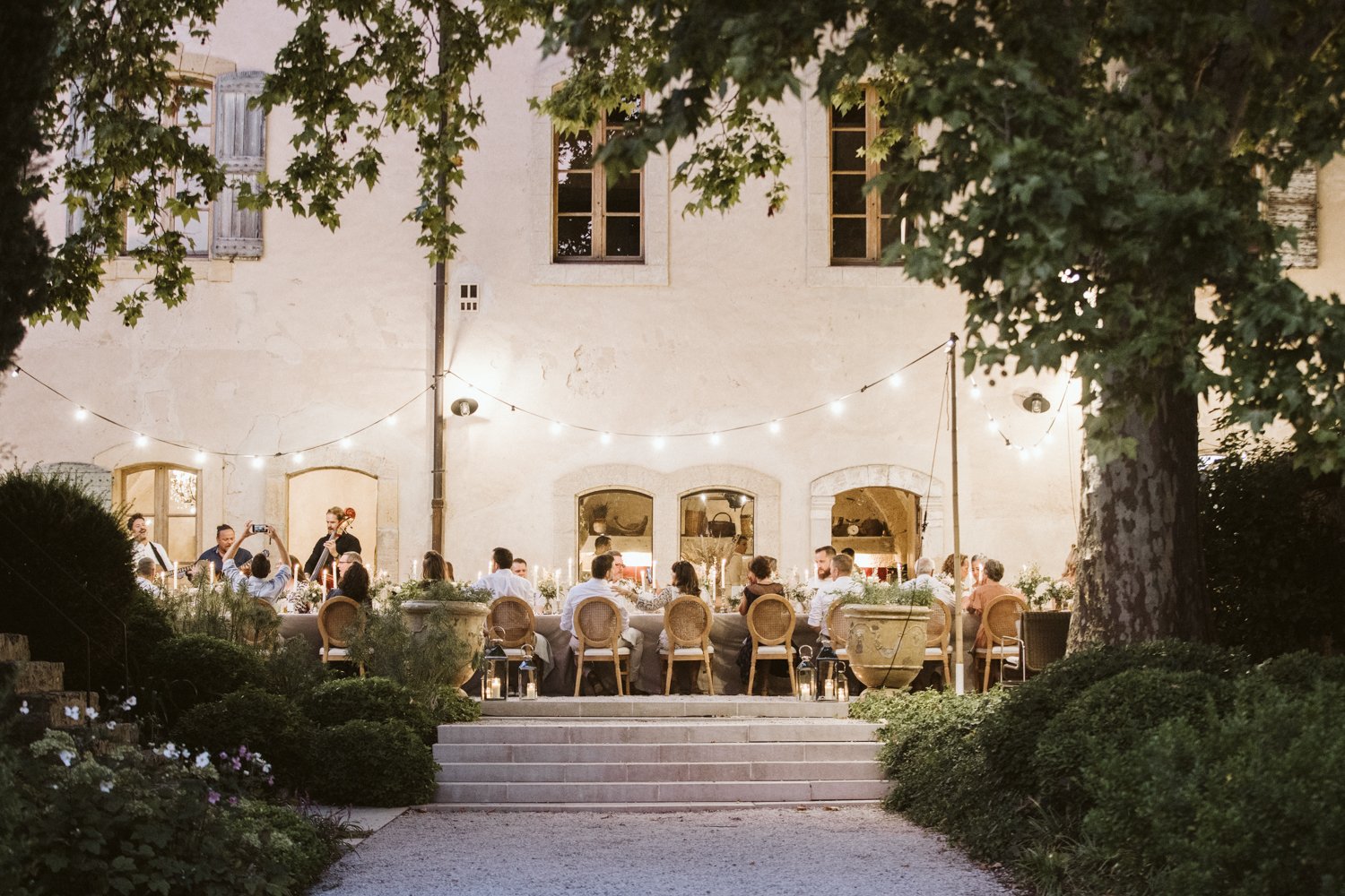 Outdoor dinner party set up on a terrace in front of an old building with string lights overhead. Guests are seated at a long table decorated with flowers, with a band playing music on the left side.