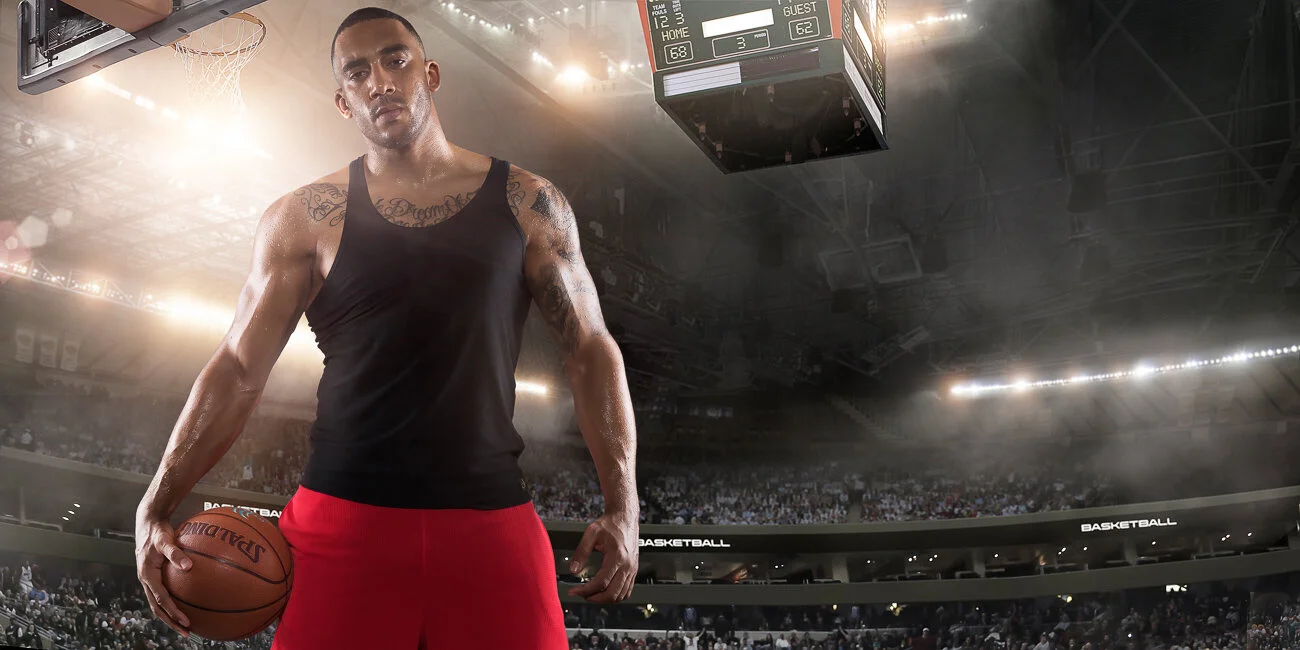  Professional basketball player standing holding basketball on an indoor floodlit basketball court full of spectators. 