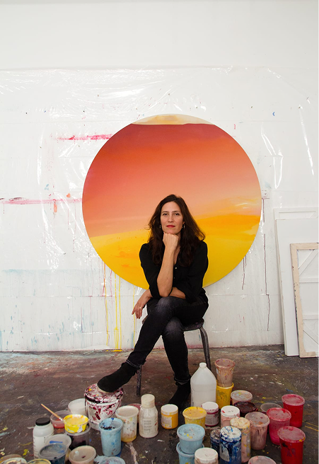 Artist Mara De Luca sits cross legged in front of her artwork, a round painting with paint materials below.