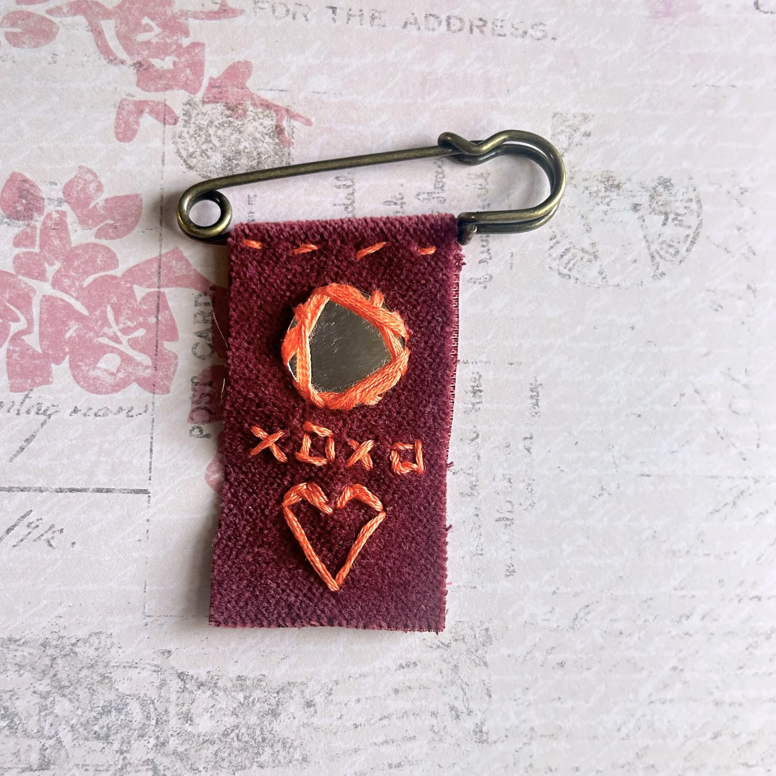 Burgundy fabric banner pin with neon thread stitches, XOXO text, heart, and a tiny mirror circle wrapped in thread on a brass-style safety pin.
