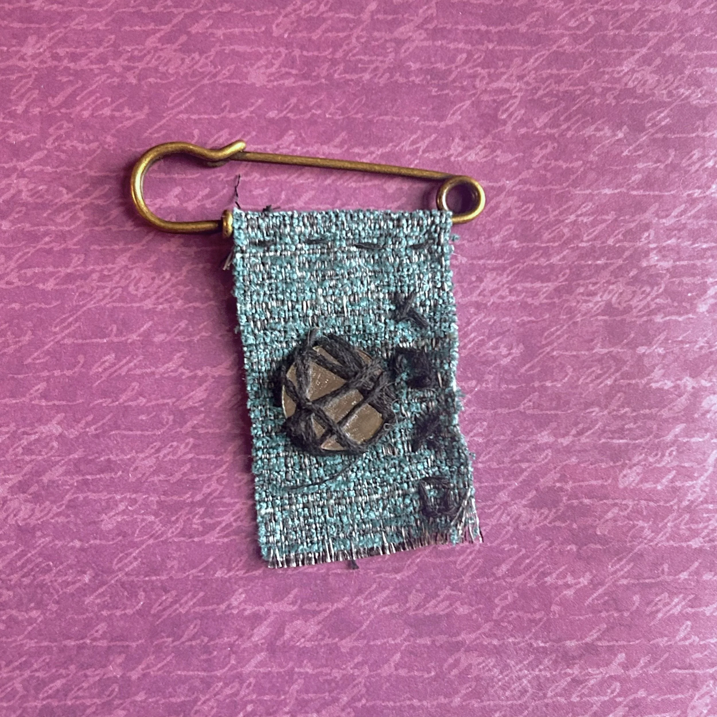 Teal fabric banner pin reading XOXO with a small mirror circle wrapped in thread on a brass-style safety pin.