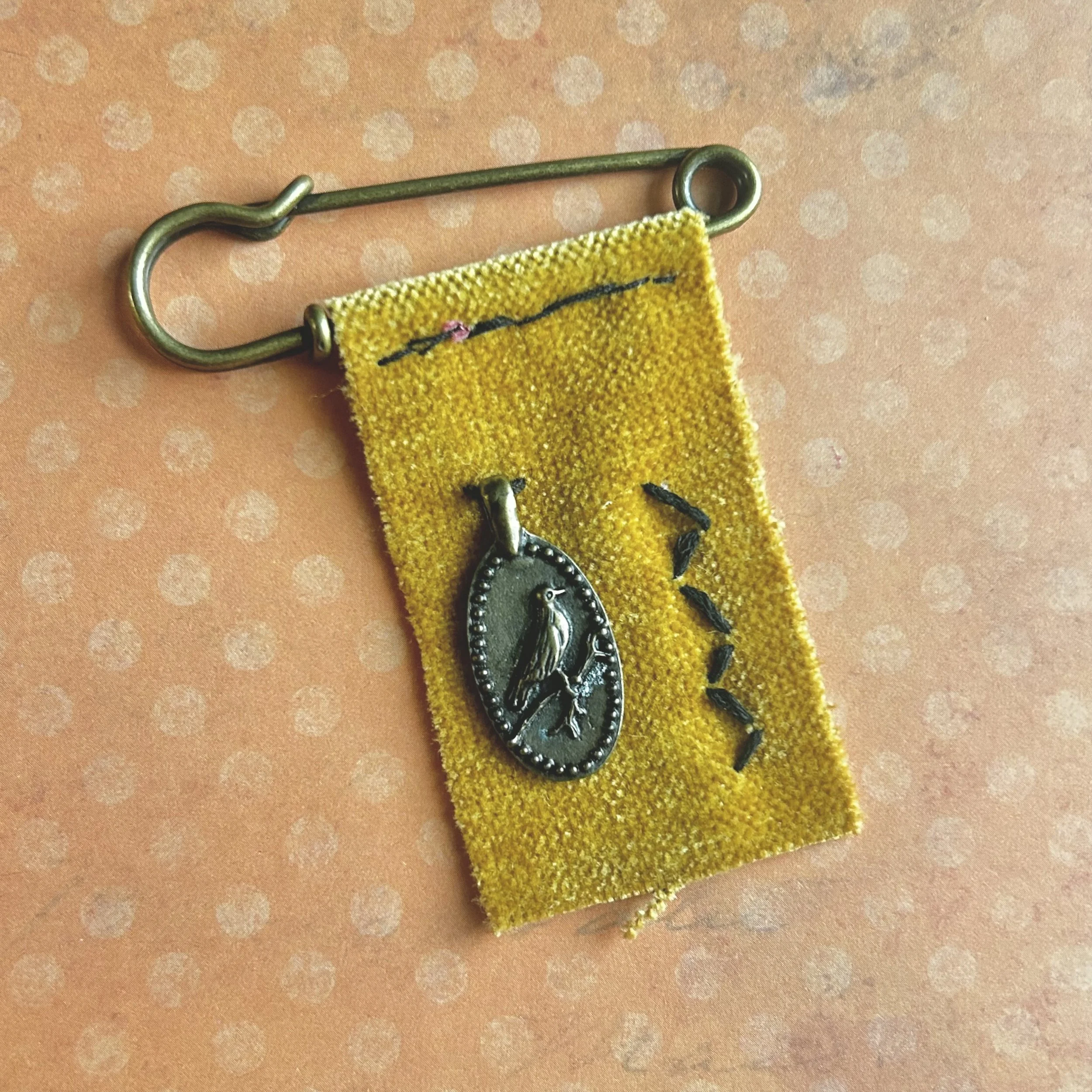 Hand-stitched yellow fabric banner pin on a brass safety pin with metal bird charm and decorative stitches.