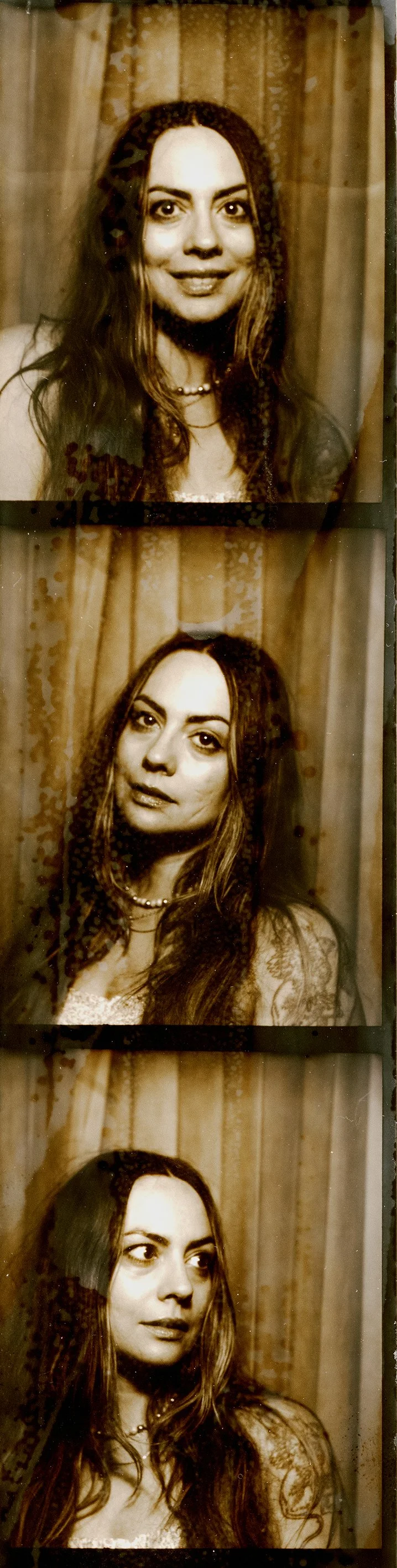 Photograph of a woman with long hair, wearing a necklace, in front of a curtain, taken in sepia tone.