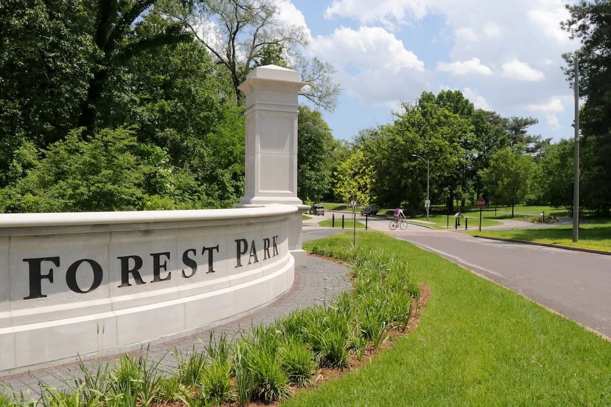 A stone marker with "Forest Park" etched marks the Wells Drive entrance to the Park as a bicyclist rides by in the background