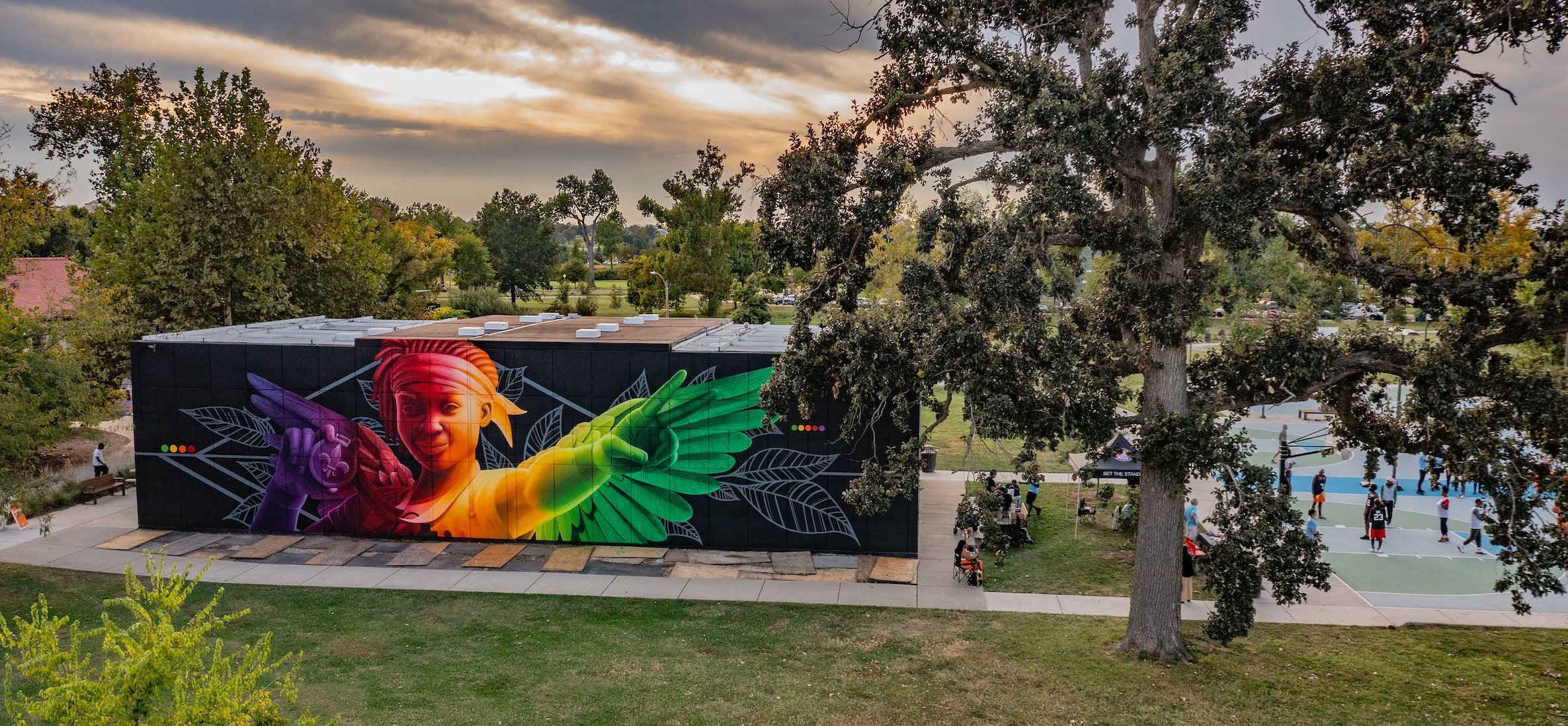 A modern interpretation of Nike as a youth, with a black background on the east wall of the racquetball courts. Green trees and a dusk sky are in the background above the courts building.