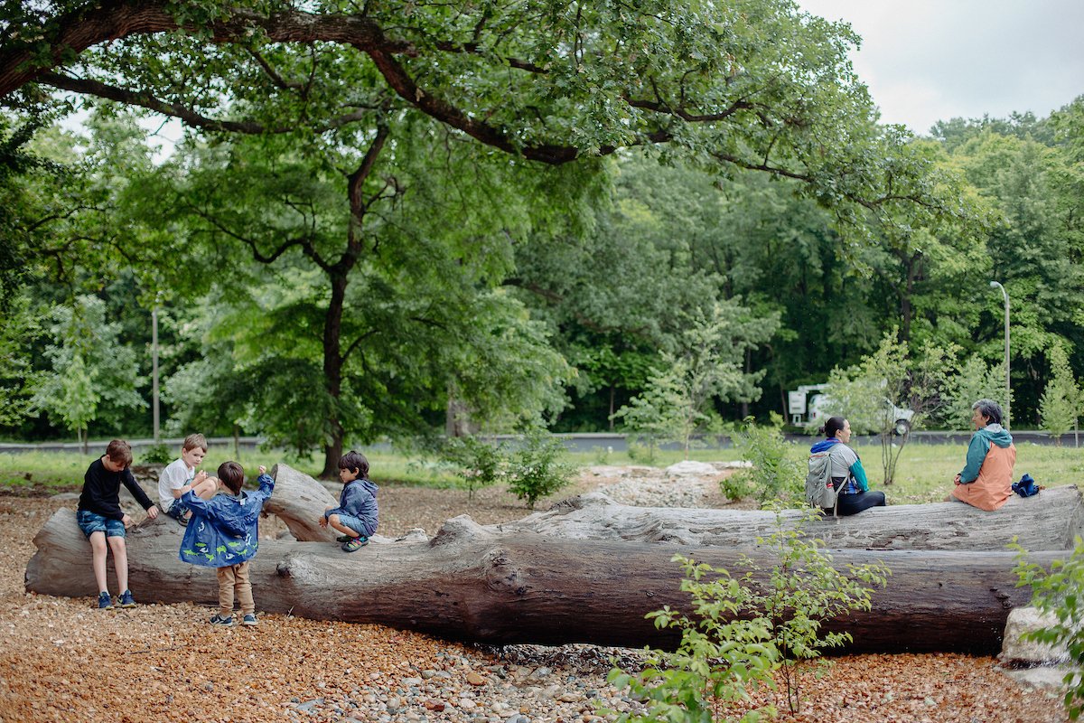 Four children play on a log in the new Nature Playscape as two adults chat nearby