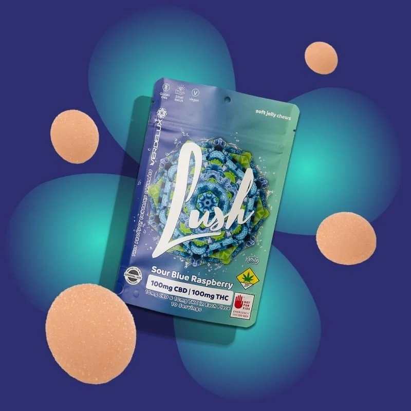 Packaging of a cannabis edible product named 'Lush' with a flavor called 'Sour Blue Raspberry', containing 100mg CBD and 100mg THC, surrounded by peach-colored gummy chews and glowing blue and teal circles in the background.