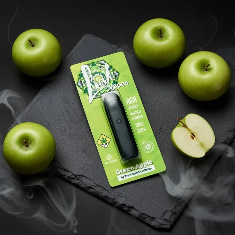 A THC vaping device on a green packaging labeled Lush Vape Green Apple, surrounded by green apples and one cut in half, on a black slate surface with wisps of smoke.