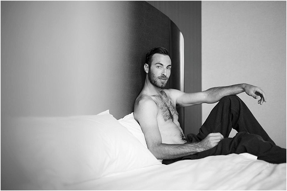 looking into the camera and shirtless a man sits on a bed
