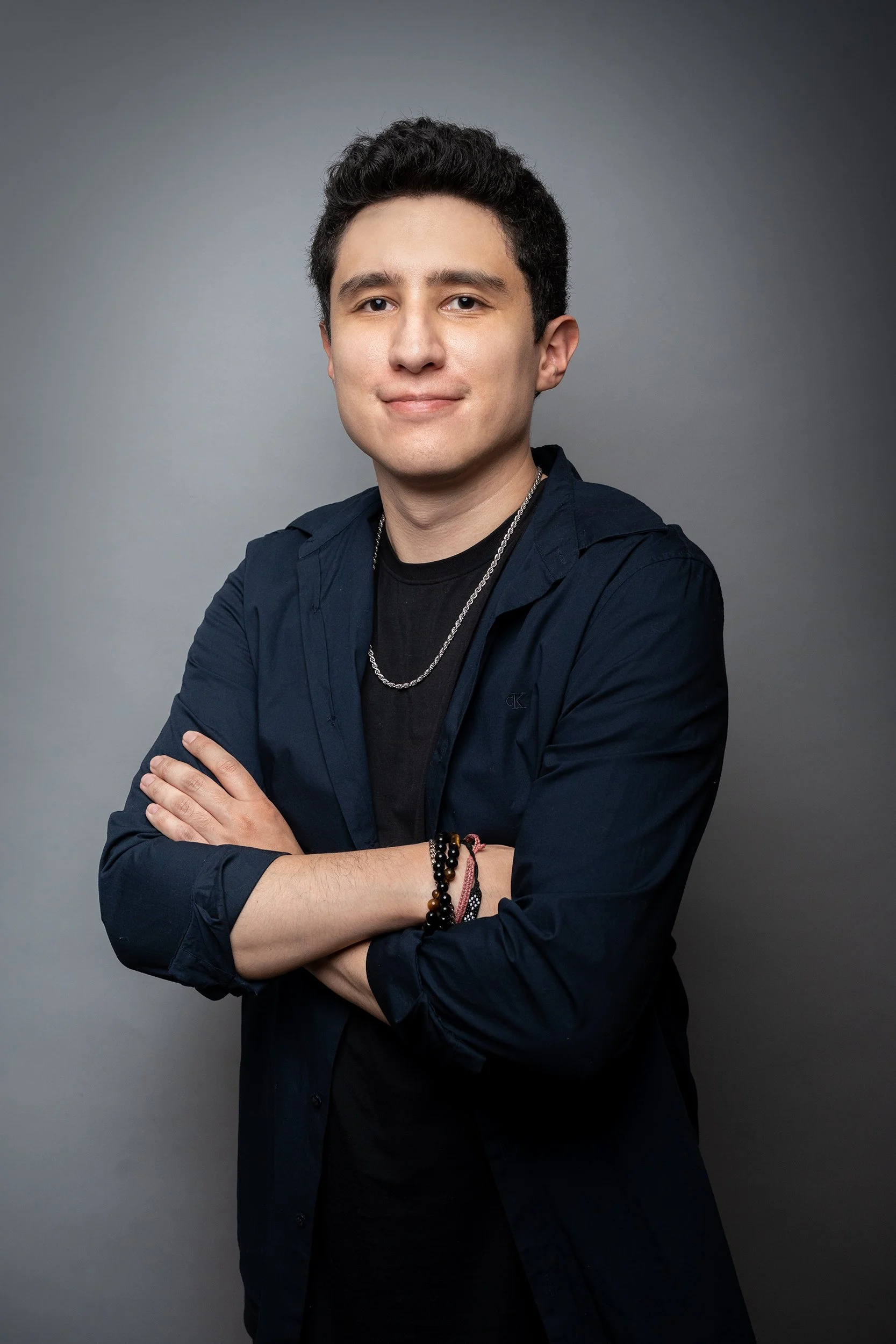 Joven hombre de cabello oscuro y sonrisa, con brazos cruzados, lleva camiseta negra y chaqueta azul oscuro, accesorios como pulseras y cadena, fondo gris oscuro.
