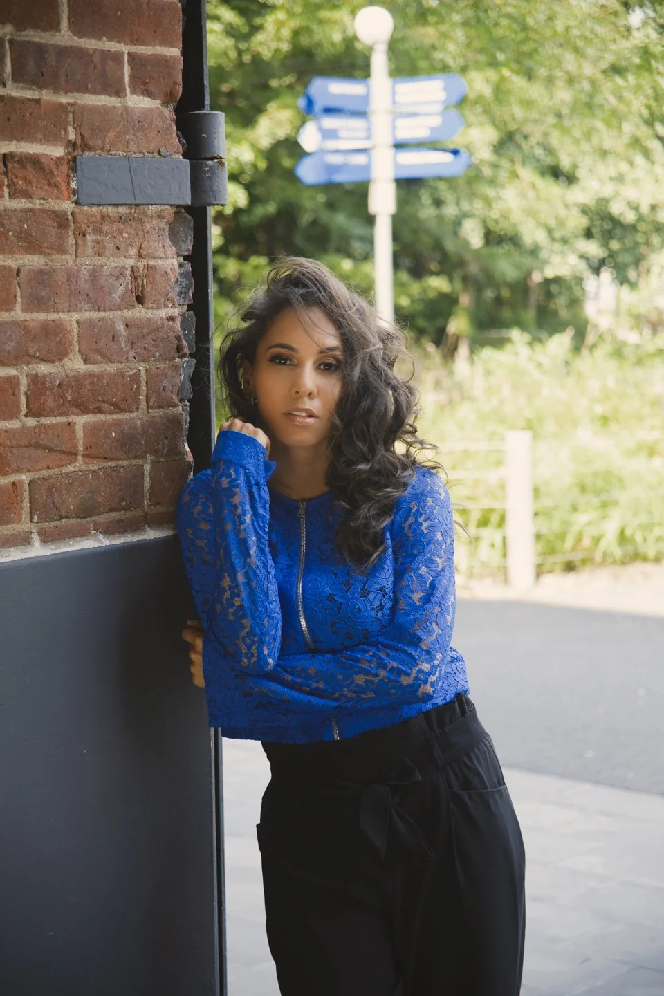 A woman with curly dark hair wearing a blue lace jacket and black pants leaning against a brick wall outdoors.