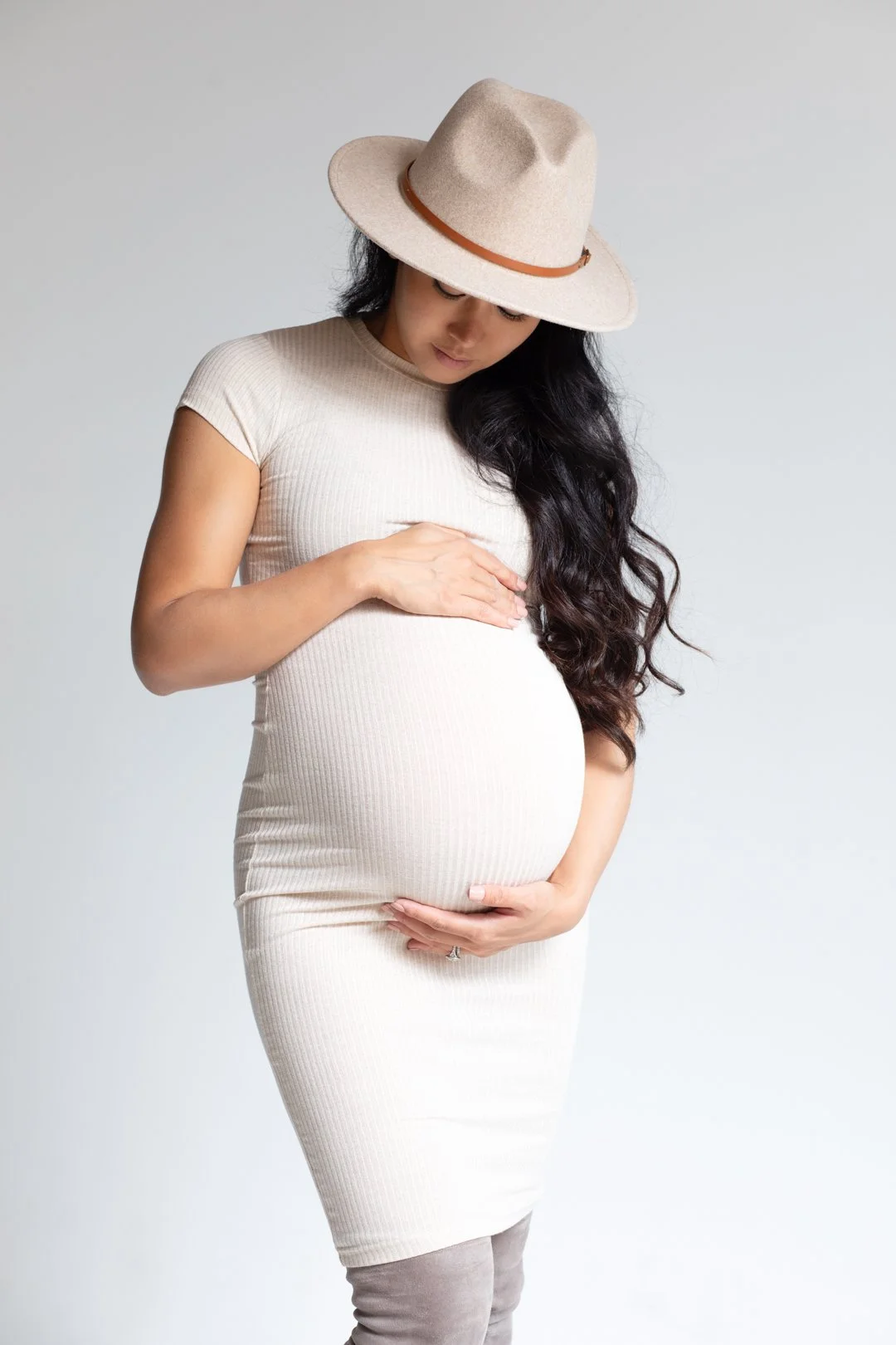 Pregnant woman in a white dress and hat, holding her belly against a plain background.