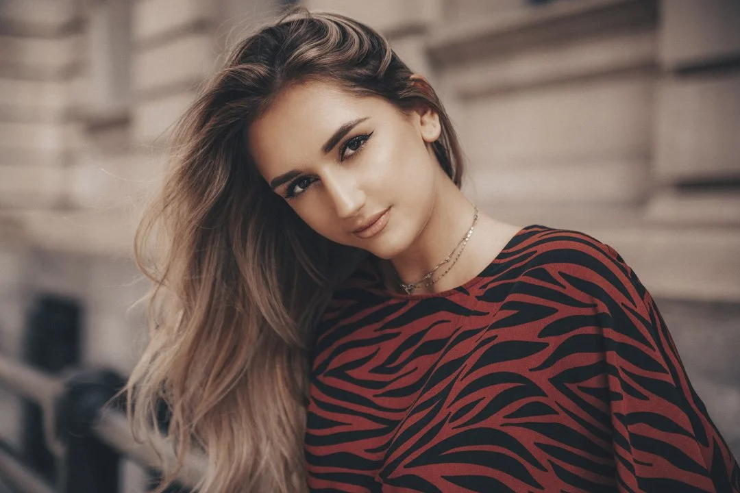 A young woman with long wavy hair, wearing a red and black patterned top, is smiling and looking at the camera with a blurred outdoor background.