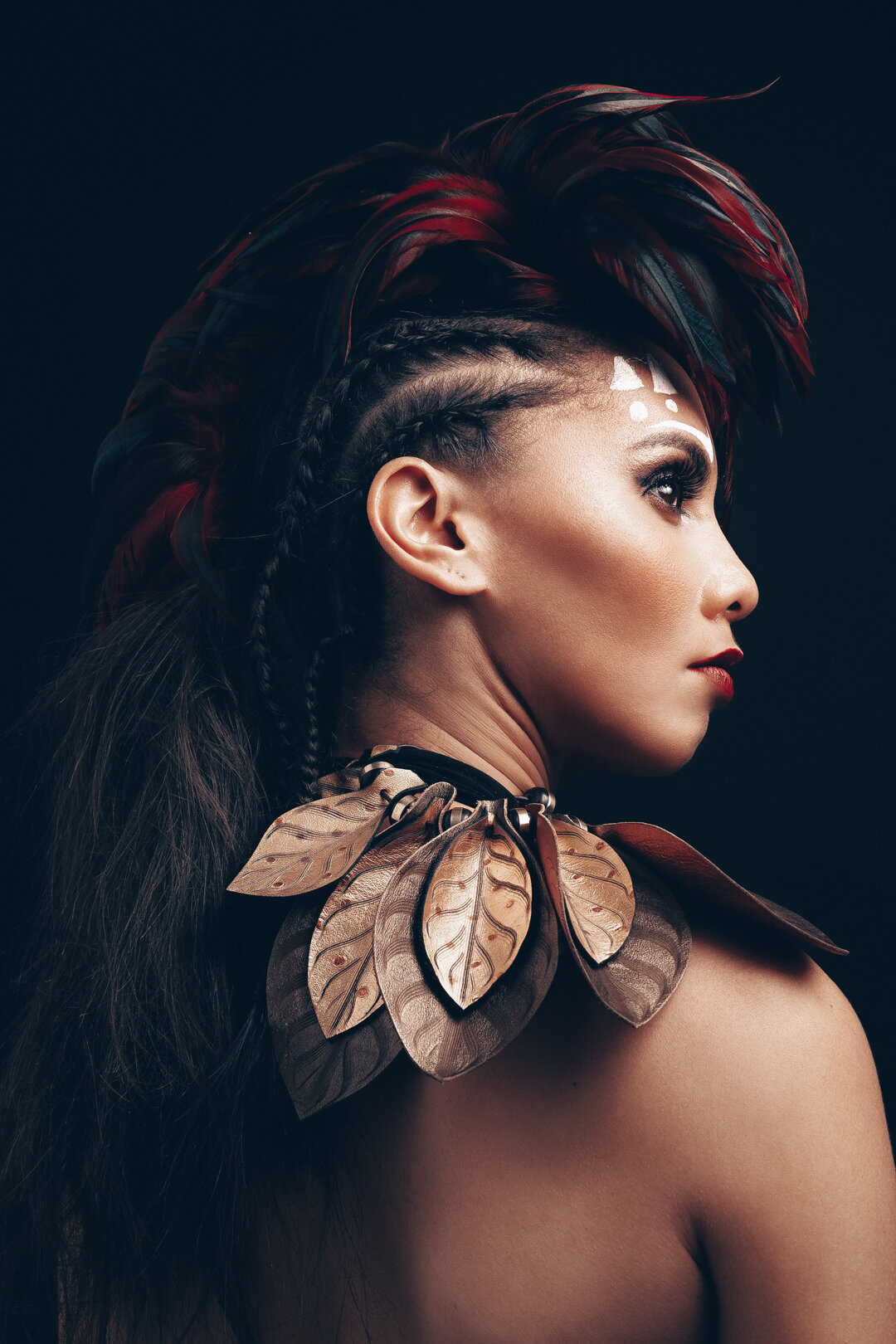 Profile of a person with an elaborate feather headdress, braided hair, and ornamental leaf necklace on a dark background.