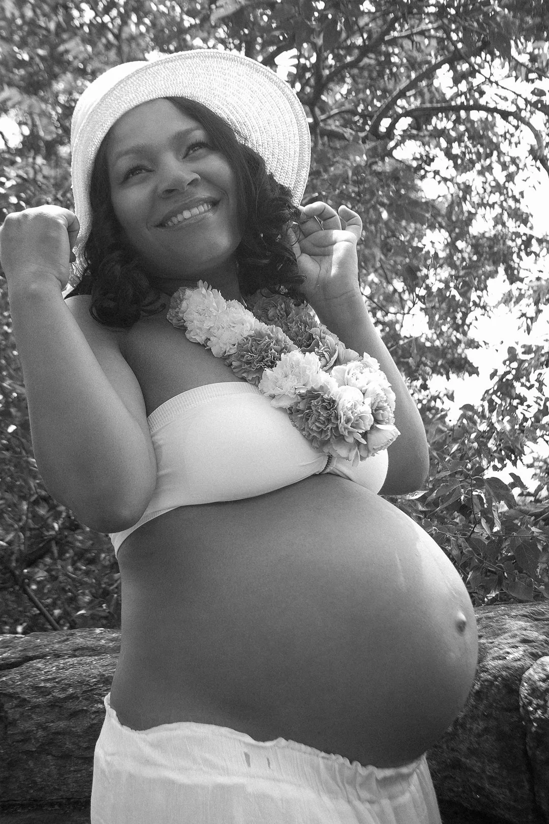 Smiling pregnant woman wearing a flower lei and a white hat, outdoors.