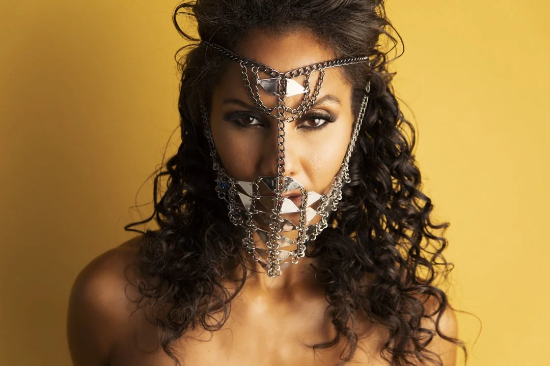 A woman with curly hair wearing an intricate face jewelry piece made of silver chains and metallic triangles, posed against a yellow background.
