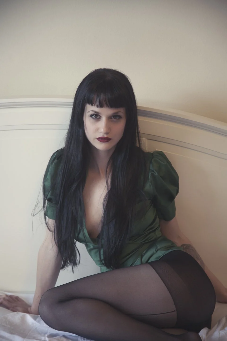 A woman with long black hair, dark lipstick, and a gothic style, sitting on a bed in a green satin dress and black stockings, looking at the camera.