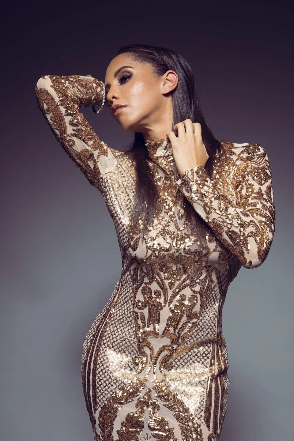 A woman in a form-fitting, long-sleeved, sheer, gold and beige dress with intricate floral and geometric patterns posed with her hand near her face against a neutral background.