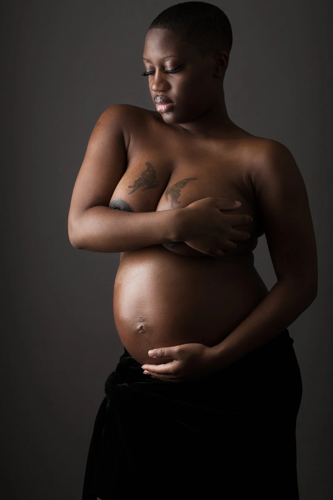 Pregnant woman with short hair, covering chest with one arm and cradling her belly with the other, against dark background.