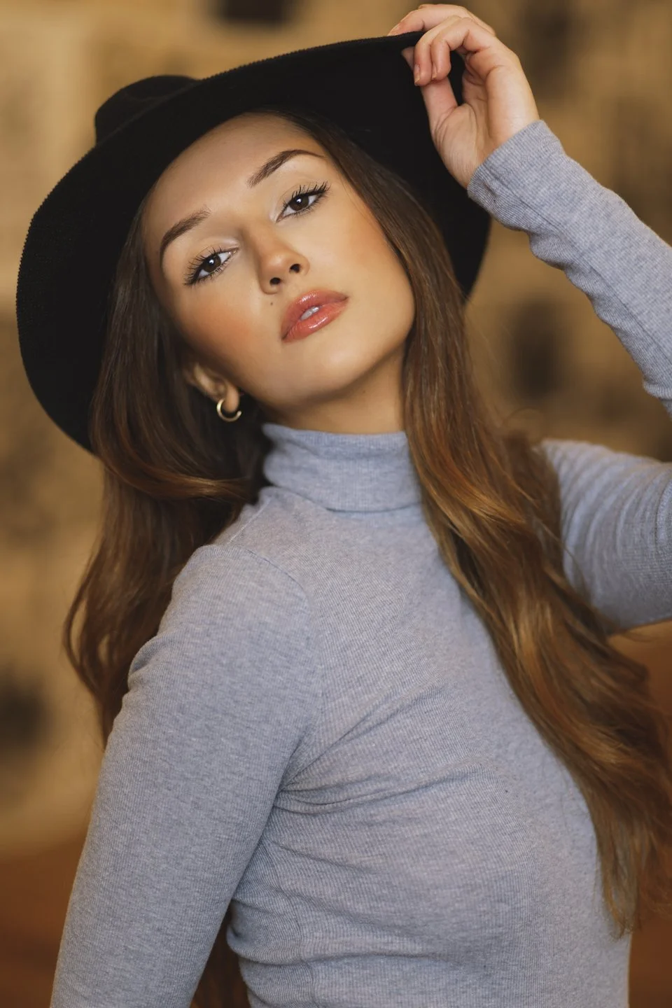 A young woman with long brown hair, wearing a black wide-brimmed hat, grey turtleneck, and a grey long-sleeve shirt, posing with her hand on her hat.
