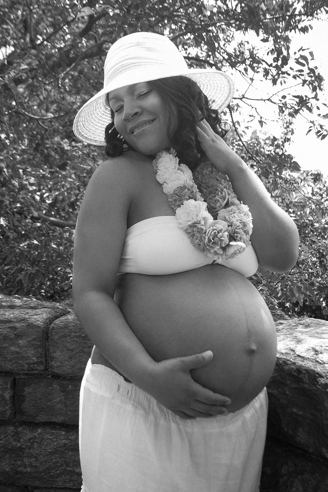 Pregnant woman wearing a hat and floral lei, smiling, outdoors.
