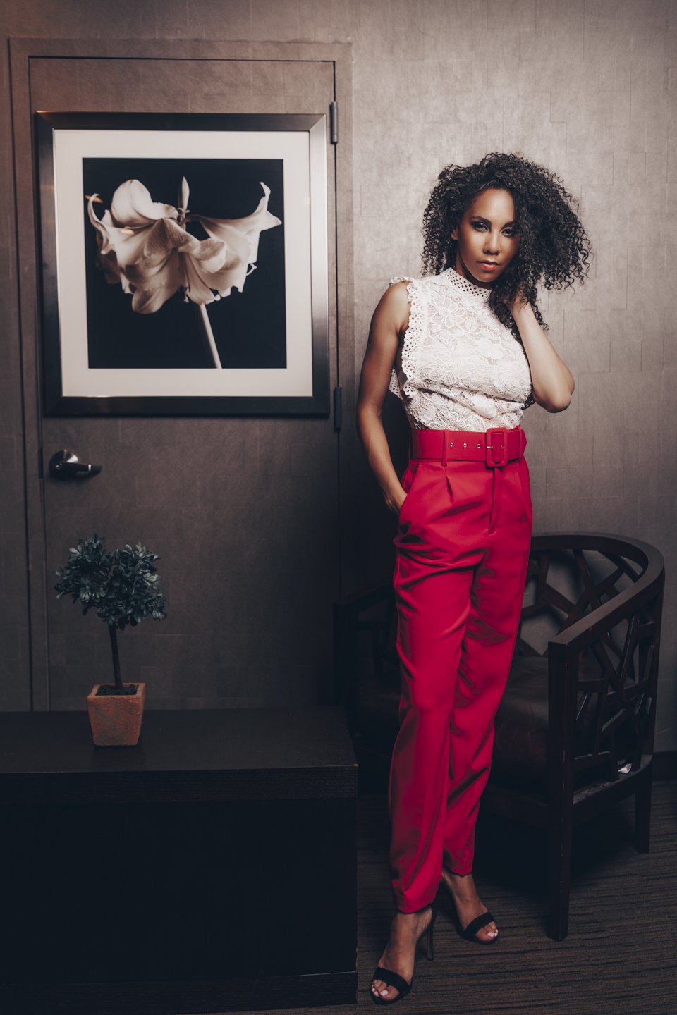 A woman with curly hair in a white lace sleeveless top and red high-waisted pants stands in an art gallery, posing next to a framed black and white photograph of a butterfly.
