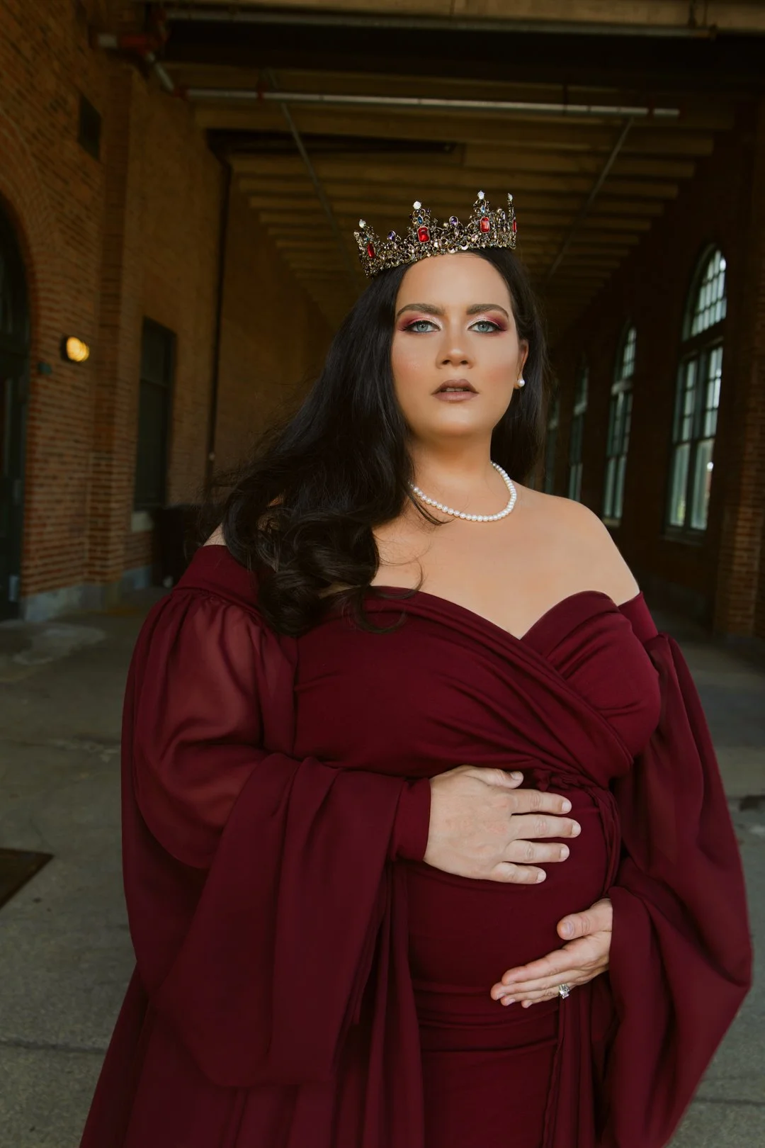 Pregnant woman wearing a dark red dress and crown, standing indoors with a hand on her belly.