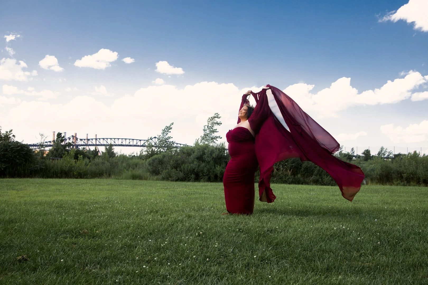 Person in a flowing burgundy dress standing on grass with arms raised, trees and a bridge in the background, under a partly cloudy sky.