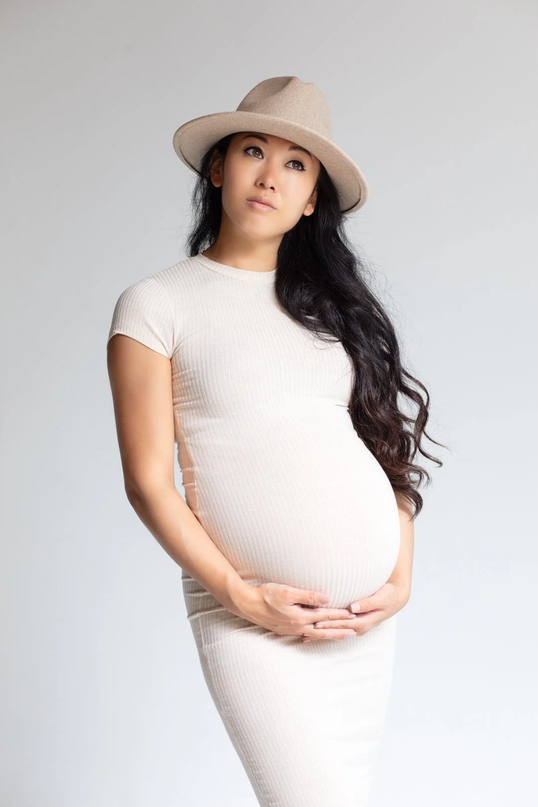 Pregnant woman in a beige dress and hat, holding her belly.